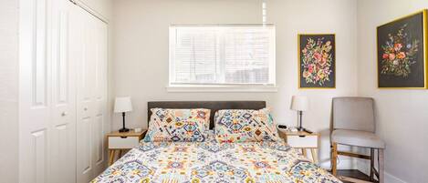 A bedroom with a bed, two bedside tables, a chair, a window with blinds, and two framed floral paintings on the wall.