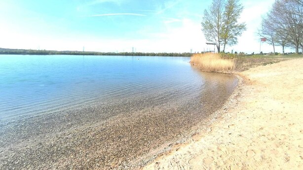 A calm lake with a pebbly shore, a clear sky, and sparse trees. - Apartment D&Y upper floor (barrier-free) (Ubstadt-Weiher)