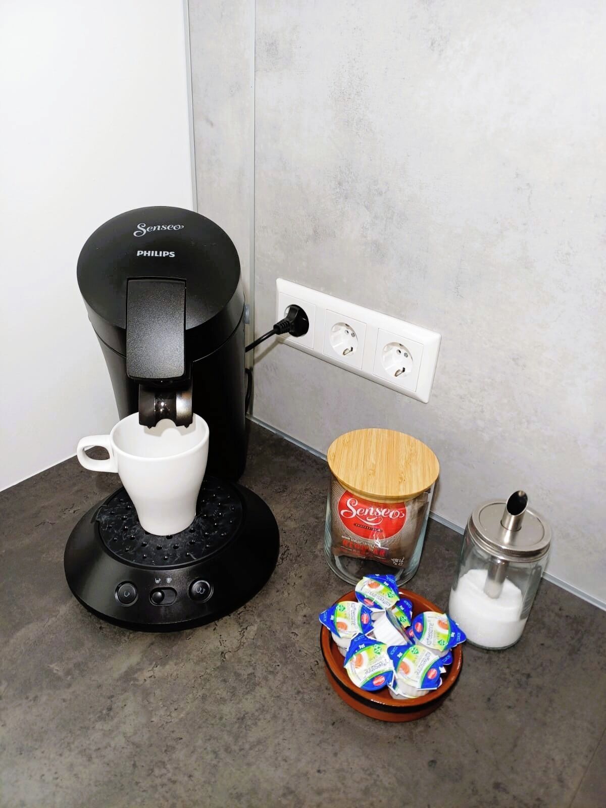 A coffee maker, a jar of coffee, and a bowl of yogurt on a countertop.