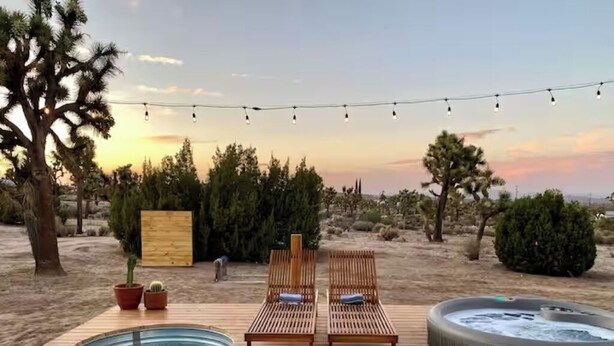 Un pontile di legno con due poltrone relax, una vasca idromassaggio e luci decorative a stringa, sullo sfondo di un paesaggio desertico.