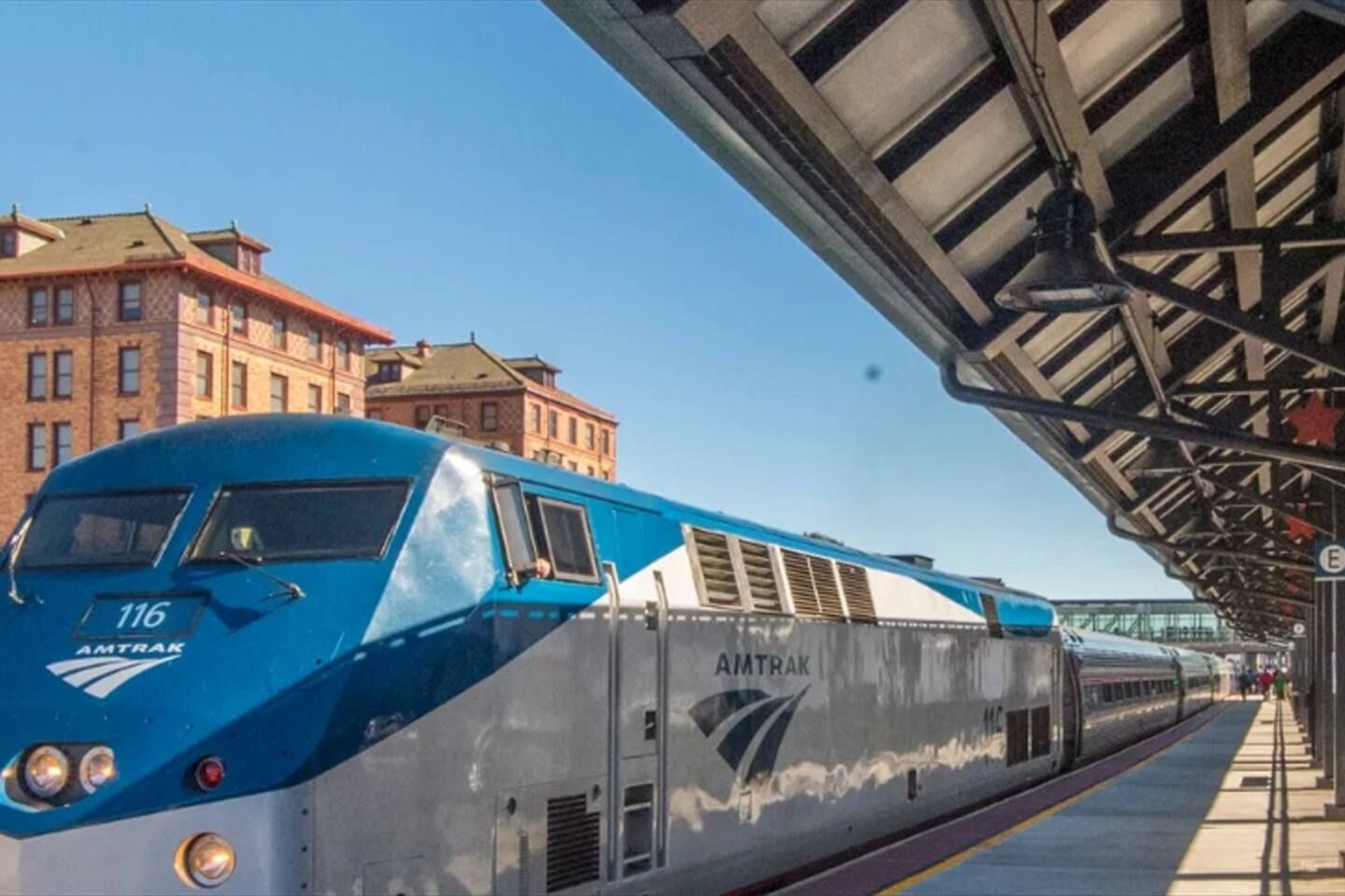 A blue Amtrak train at a station with a covered platform and buildings in the background.