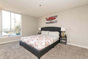 A bedroom with a large window, a bed with a floral bedspread, a black headboard, and a small bedside table with a lamp. - Hollywood 2BR Suite with Rooftop Pool & Cityscape (Los Angeles)
