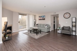 A spacious living room with a large window, a gray sofa, a coffee table, and a fireplace. - Hollywood 2BR w/ Rooftop Pool & City Views (Los Angeles)