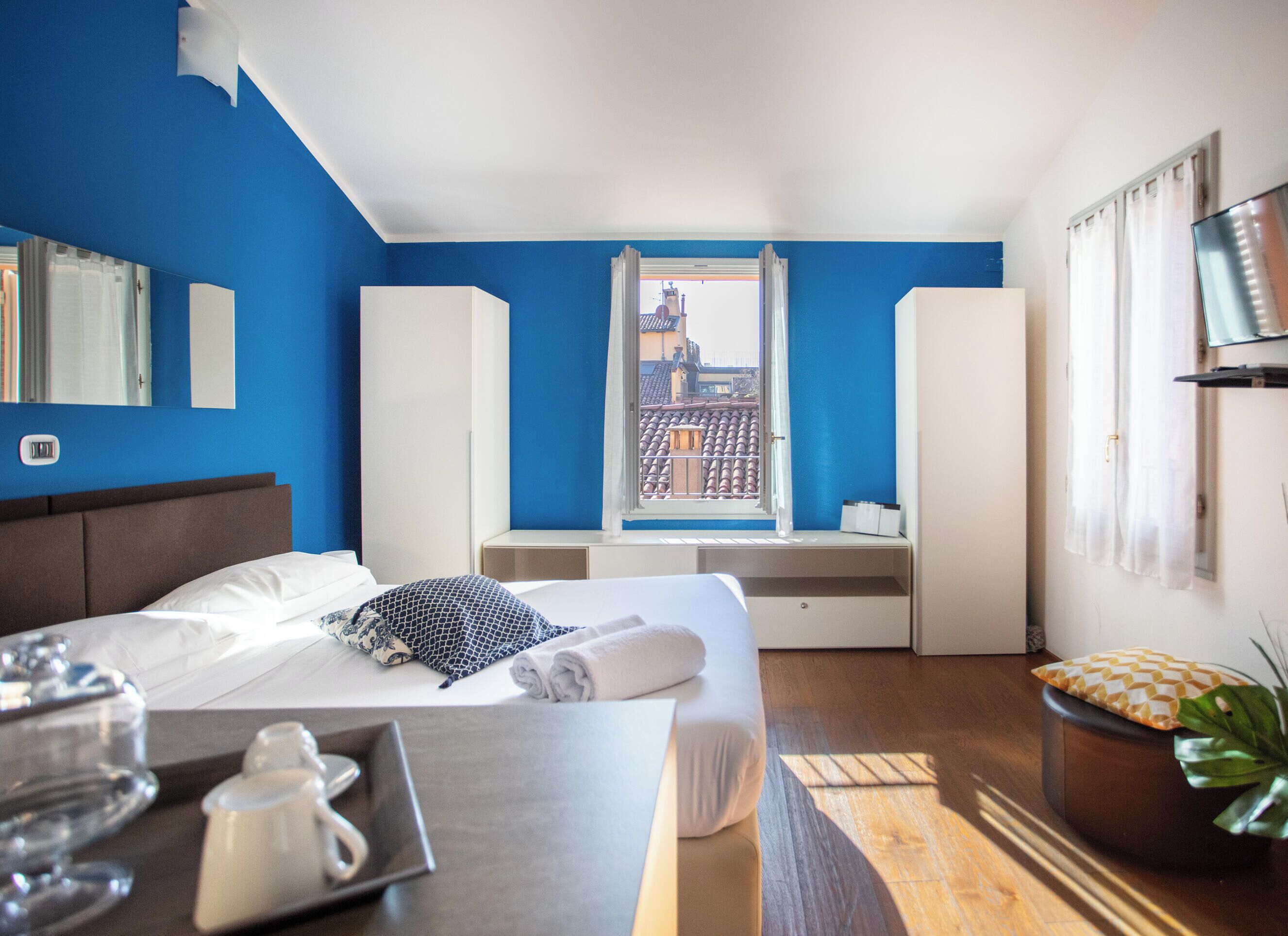 Une chambre d’hôtel avec un lit, une fenêtre donnant sur des immeubles, un bureau avec une plante et un miroir au mur.