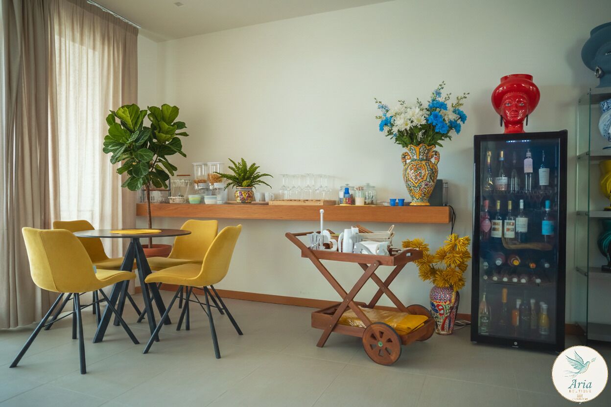 A modern dining area with yellow chairs, a wooden table, a small cart with kitchen items, and a refrigerator with bottles.