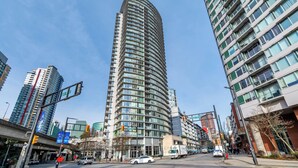 A tall, curved building with multiple floors and large windows, surrounded by other high-rise buildings and a busy street with traffic lights and vehicles. - Bright 3BR Downtown Vancouver Condo w/ Pool & Gym (Vancouver)
