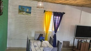 Un coin repas avec un plafond en bois, une table recouverte d’une nappe blanche et des chaises avec des housses à motifs. Un tableau est accroché au mur et un téléviseur est posé sur un meuble.