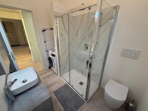 Une salle de bain moderne dotée d’une douche à parois de verre, de toilettes et d’un lave-linge dans la pièce adjacente.