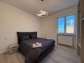Une chambre moderne avec un grand lit, une table de chevet, un radiateur et une fenêtre donnant sur des immeubles.