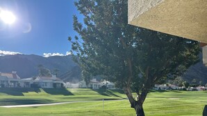 Ein gepflegter Golfplatz mit freiem Blick auf die dahinterliegenden Berge, ein Baum im Vordergrund und ein Gebäude mit Satteldach.