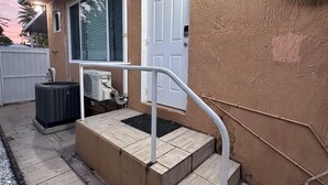 Una casa con porche cubierto, una puerta blanca y un equipo de aire acondicionado instalado en la pared exterior.