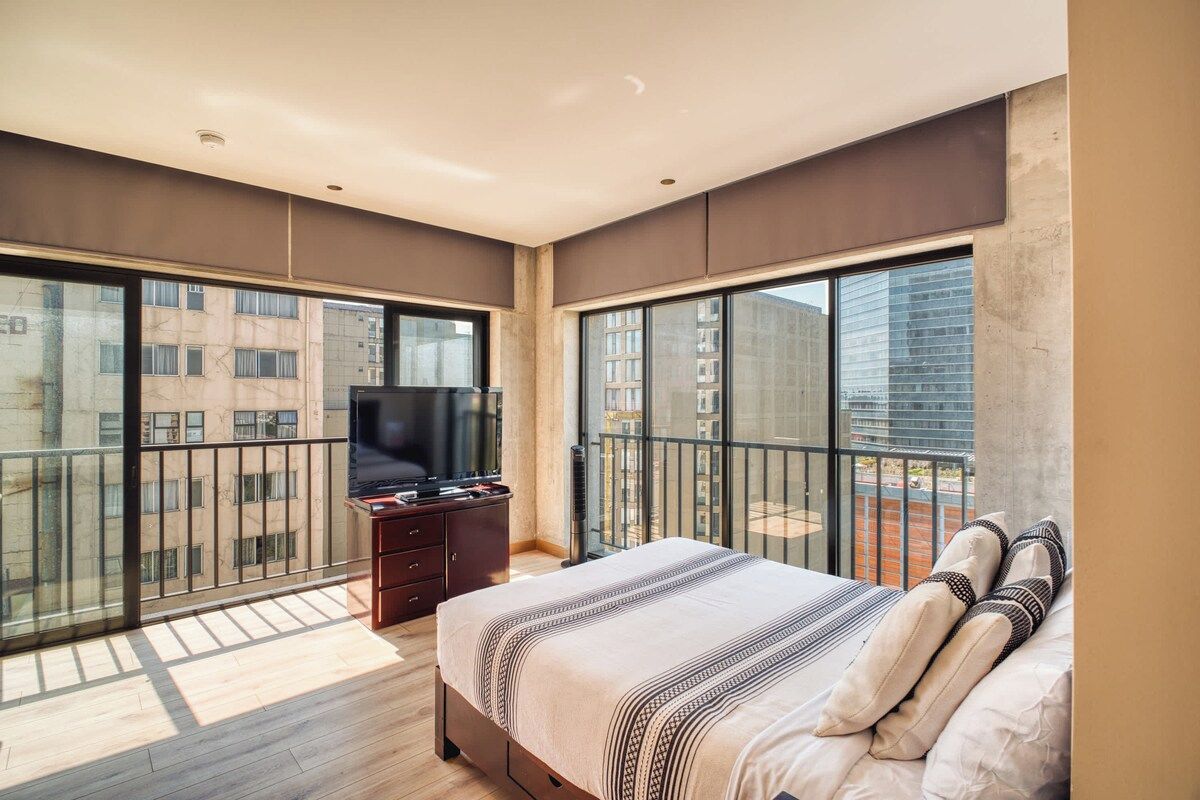Een moderne slaapkamer met een groot bed, een televisie en een balkon met uitzicht op gebouwen.