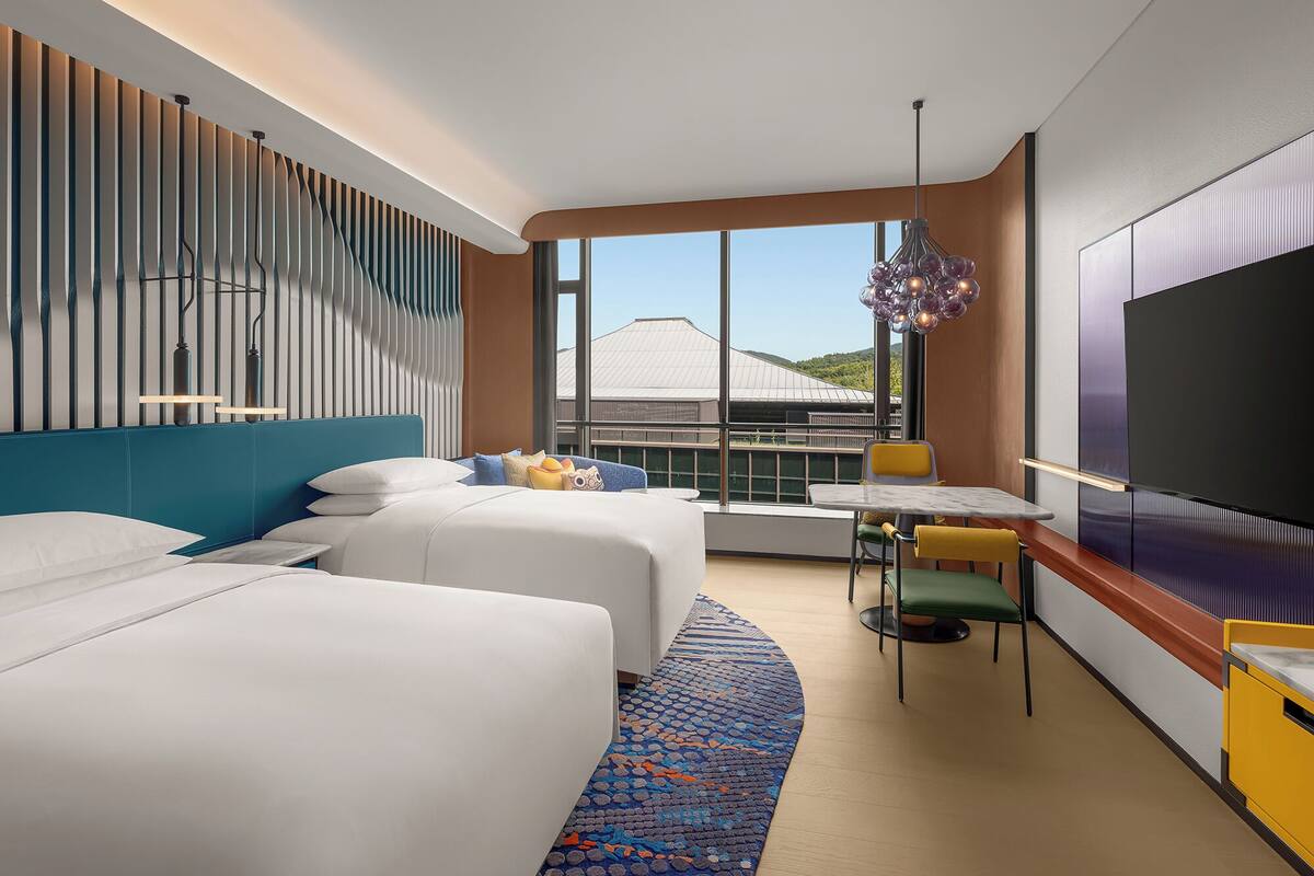 Habitació d'hotel moderna amb dos llits, zona de menjador i una finestra gran amb vista a un edifici.