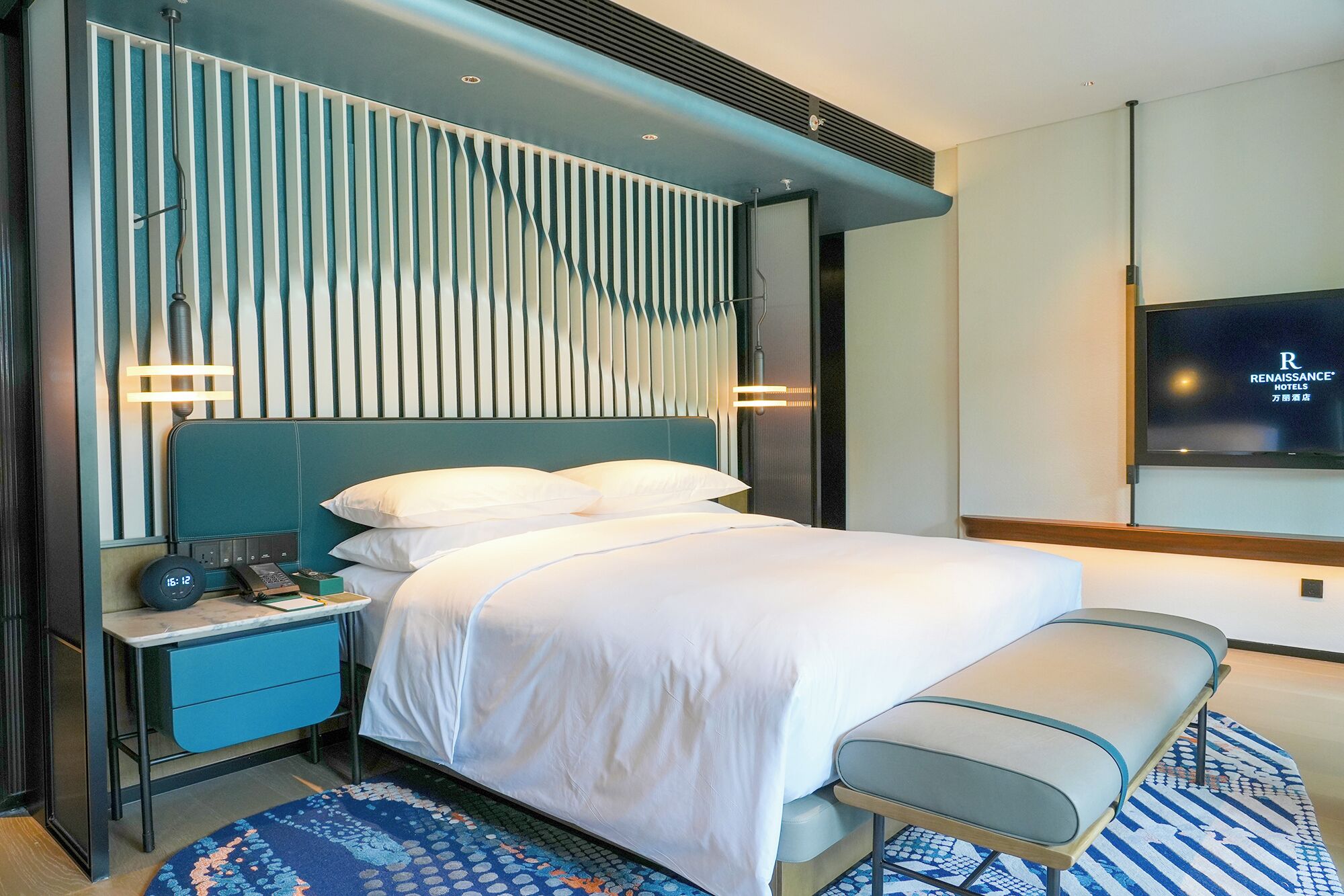 Una habitación de hotel moderna con cama, mesita de noche, banco y televisor fijado a la pared.