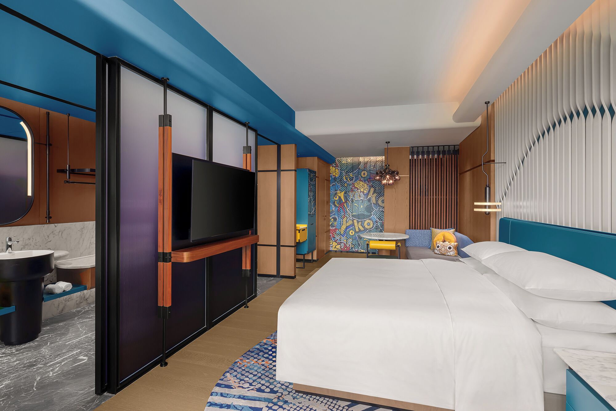 Habitación de hotel moderna con una cama grande, un televisor montado en un panel de madera, un baño con lavabo e inodoro, y un mural de pared colorido.
