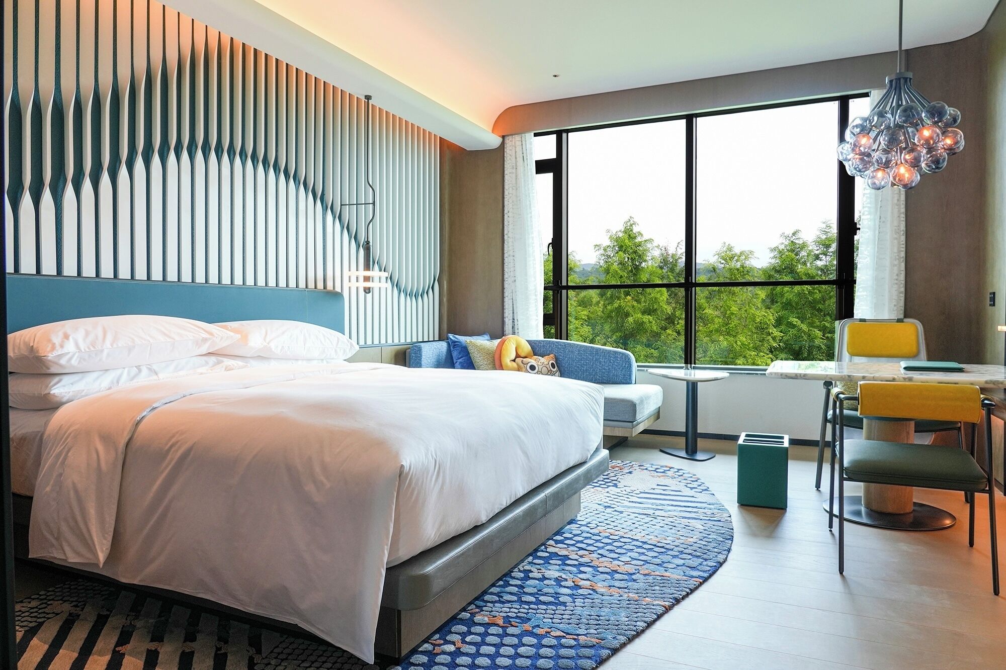 A modern bedroom with a large bed, a seating area with a table, and a view of greenery outside.