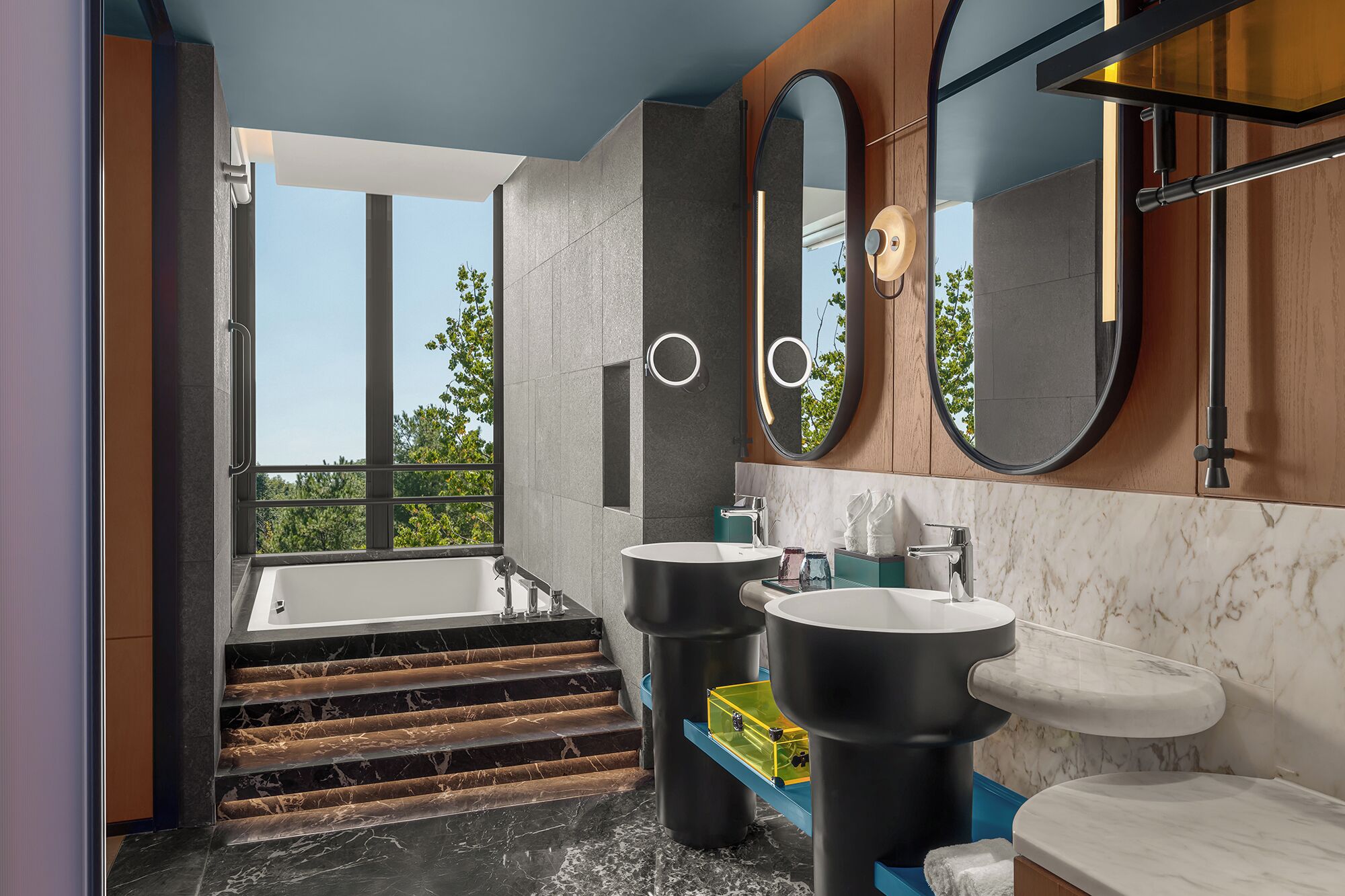 A modern bathroom with a large bathtub, two sinks, and a window offering a view of trees.