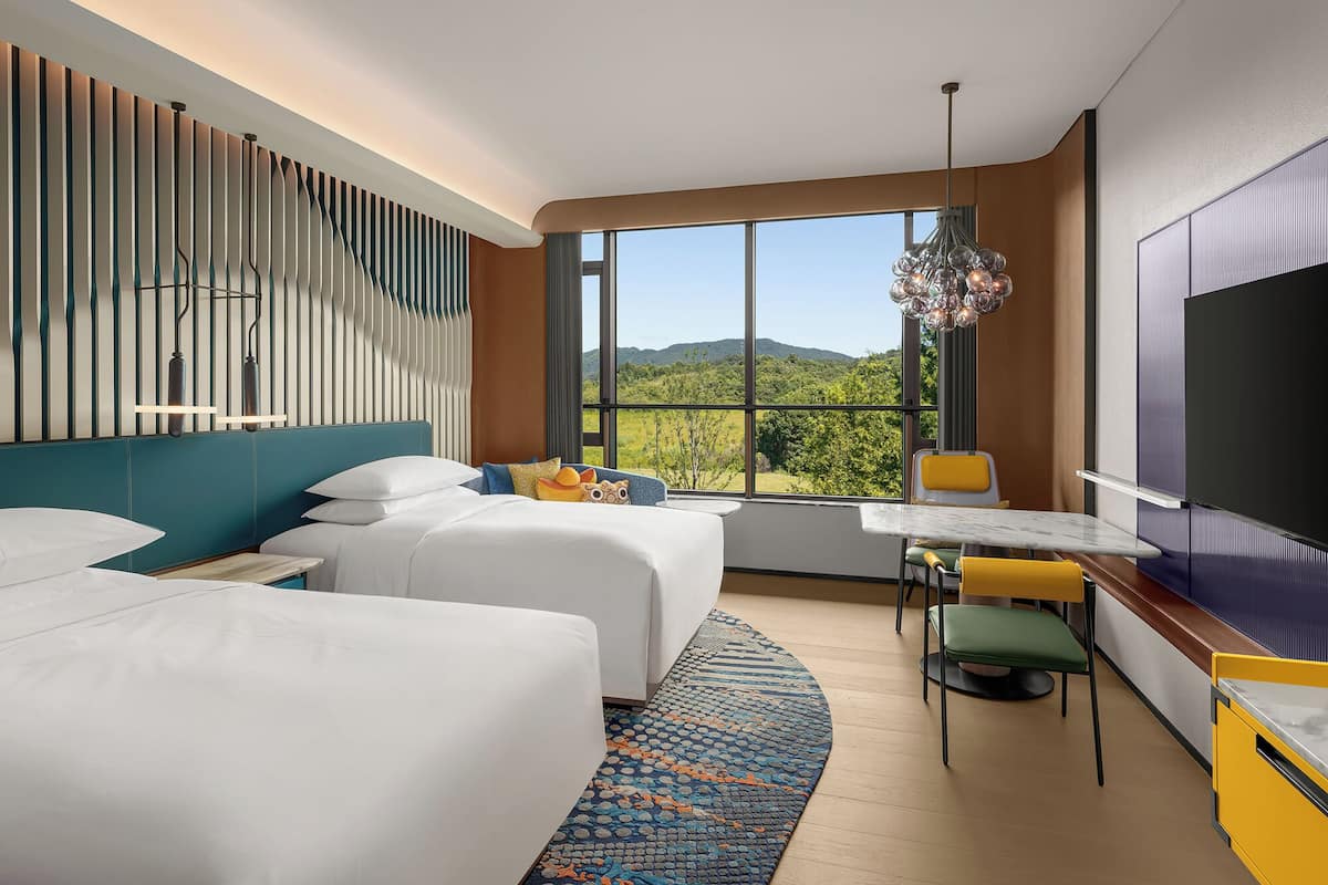 Habitació d'hotel amb dos llits, un escriptori, una cadira, un televisor i una finestra gran amb vista a la muntanya.