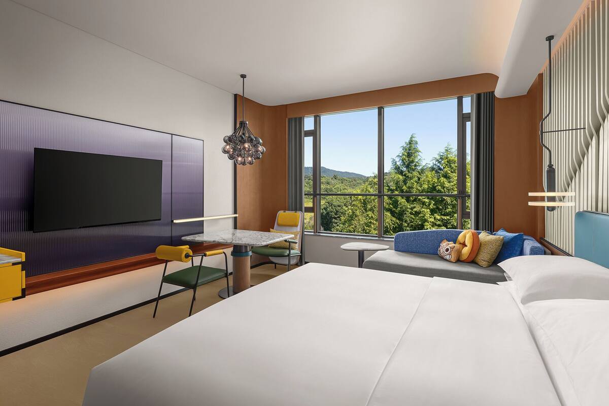 Habitació d'hotel moderna amb un llit gran, una televisió de pantalla plana, zona de menjador amb taula i cadires, i vista a l'exterior.