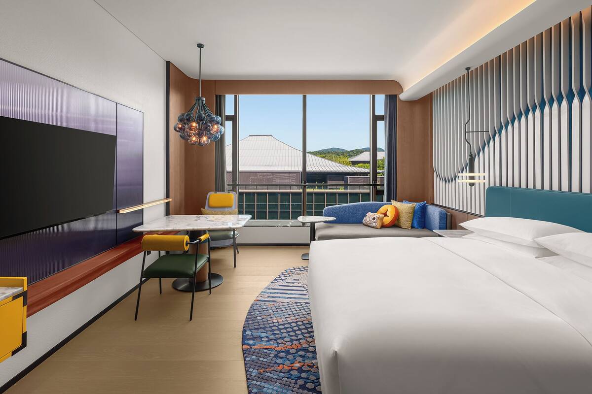 Habitació d'hotel moderna amb un llit gran, una taula de treball amb una cadira, una televisió i una finestra ampla que ofereix vistes a edificis i turons.