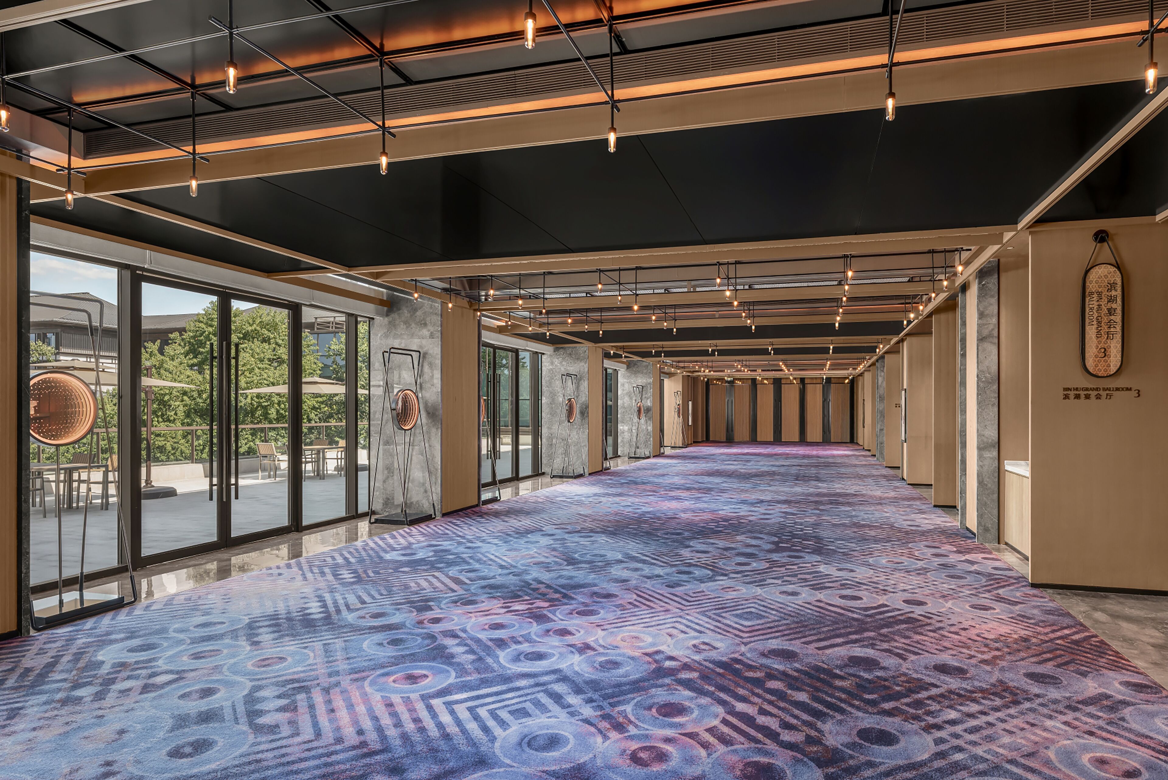 Un pasillo de hotel con alfombra estampada, ventanas amplias y luminarias modernas.