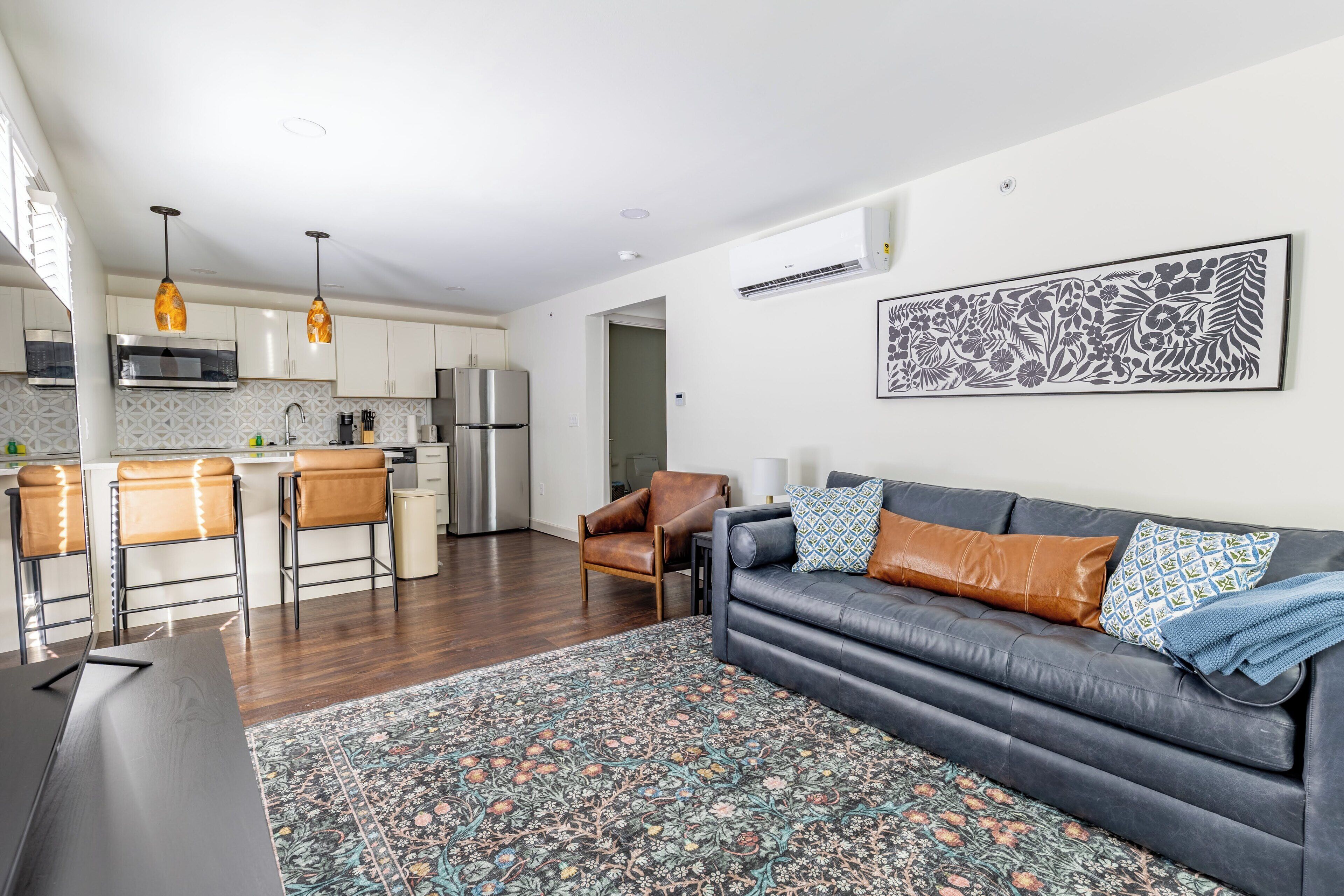 A modern kitchen with stainless steel appliances, a dining area with a dark wooden table, and a living room with a grey sofa and patterned rug.
