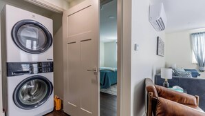 Um espaço de estar compacto com máquina de lavar frontal, uma porta para um quarto, sofá e ar condicionado fixado na parede.