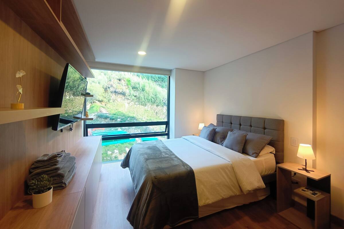 Ein modernes Schlafzimmer mit Bett, Nachttisch, Fernseher und Blick auf eine grüne Landschaft.