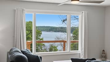 A room with a large window offering a view of a lake and forest, a ceiling fan, and a small table.