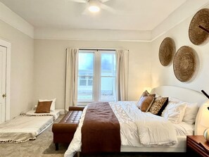 Ein Schlafzimmer mit einem großen Bett, einem kleineren Bett, einer Bank und einem Fenster mit Vorhängen.