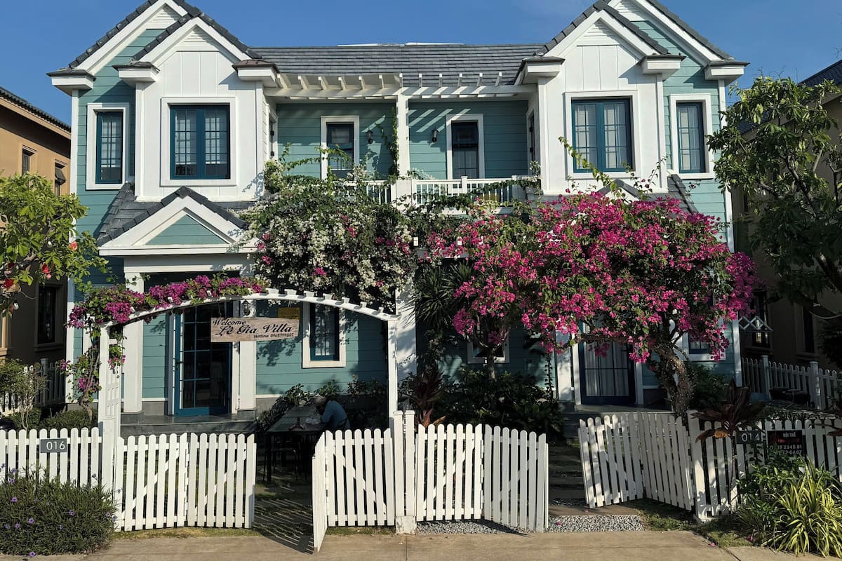 A two-story house with a white picket fence, a porch with a sign, and pink bougainvillea flowers.