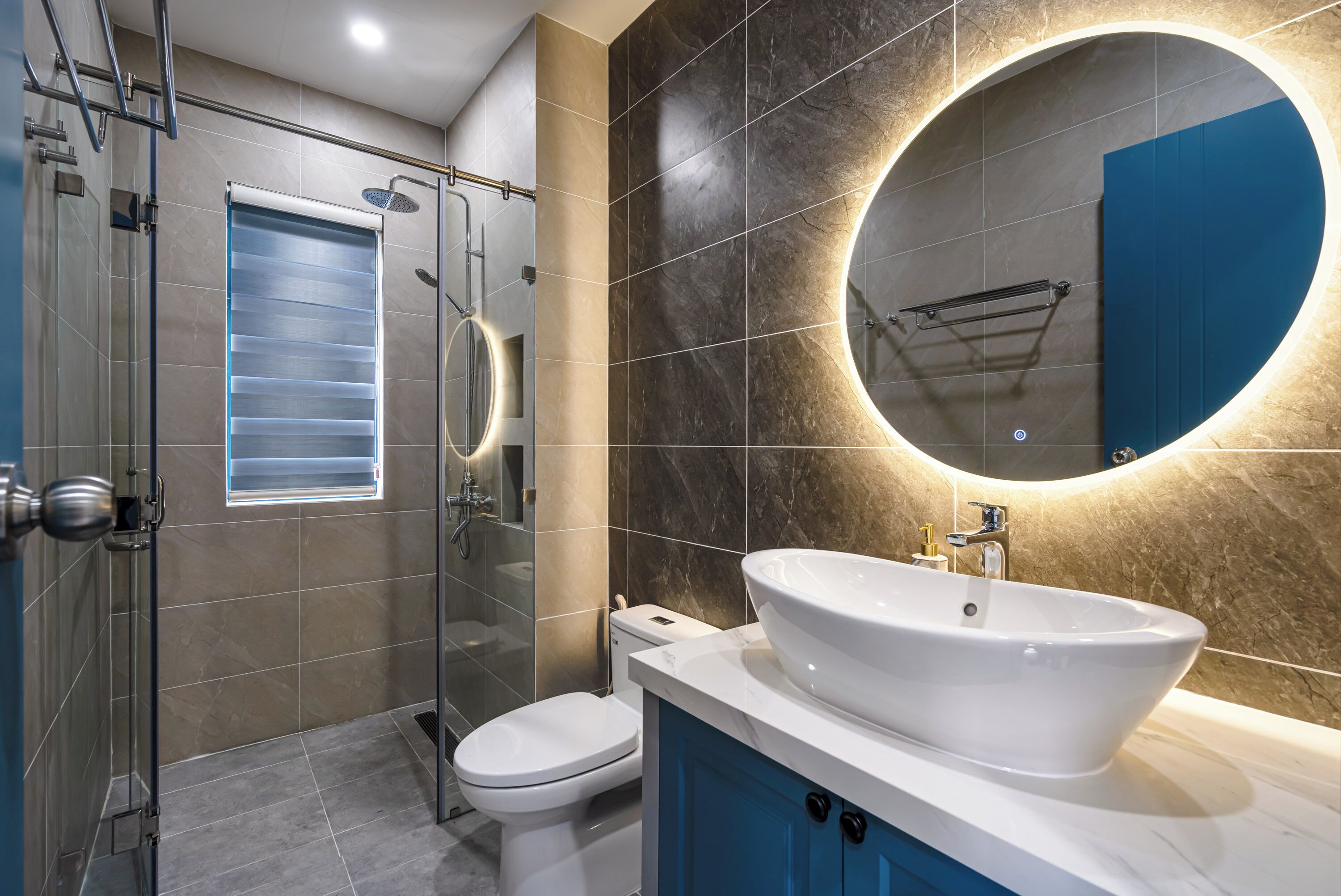 A modern bathroom with a glass shower enclosure, a toilet, a large sink with a vessel basin, and a round mirror with built-in lighting.