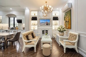 A spacious living room with a high ceiling, a chandelier, a flat-screen TV mounted above a fireplace, and a dining area with a table and chairs. - Le Gia Villa Nova World Phan Thiet (Tien Thanh)
