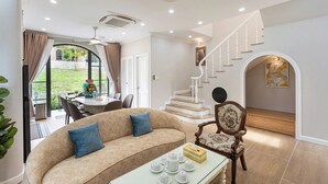 Ein Wohnzimmer mit einem beigefarbenen Sofa, einem Glastisch und einem Holzstuhl. Eine Treppe mit weißer Geländer führt zu einer oberen Etage.