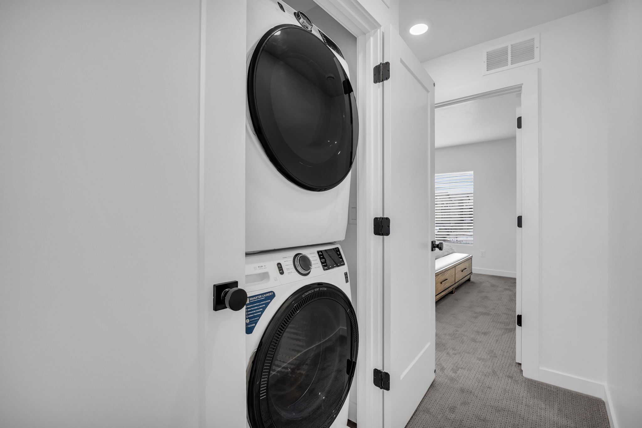 Een wasruimte met gestapelde wasmachines en drogers, een kleine slaapkamer met een bed en nachtkastje, en een gang met een vloerbedekking.