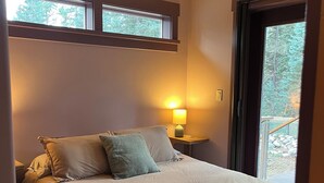 Ein Schlafzimmer mit einem Bett, Kissen, einem Nachttisch mit Lampe und einem Fenster mit Blick auf Bäume.