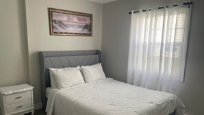 A bedroom with a bed, a nightstand, a framed picture on the wall, and a window with sheer curtains. - Your perfect getaway: stunning views, prime location, 5-Star Amenities (St. John's)