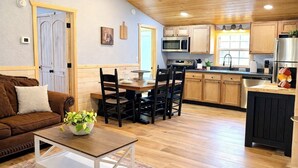 Una sala de estar acogedora con un sofá marrón, una mesa de centro con una planta y un comedor con sillas y una mesa de madera. La cocina tiene armarios de madera, un microondas y un refrigerador.