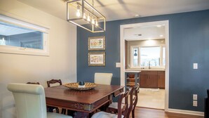 A dining area with a wooden table and chairs, framed artwork on the wall, and a view into the kitchen.