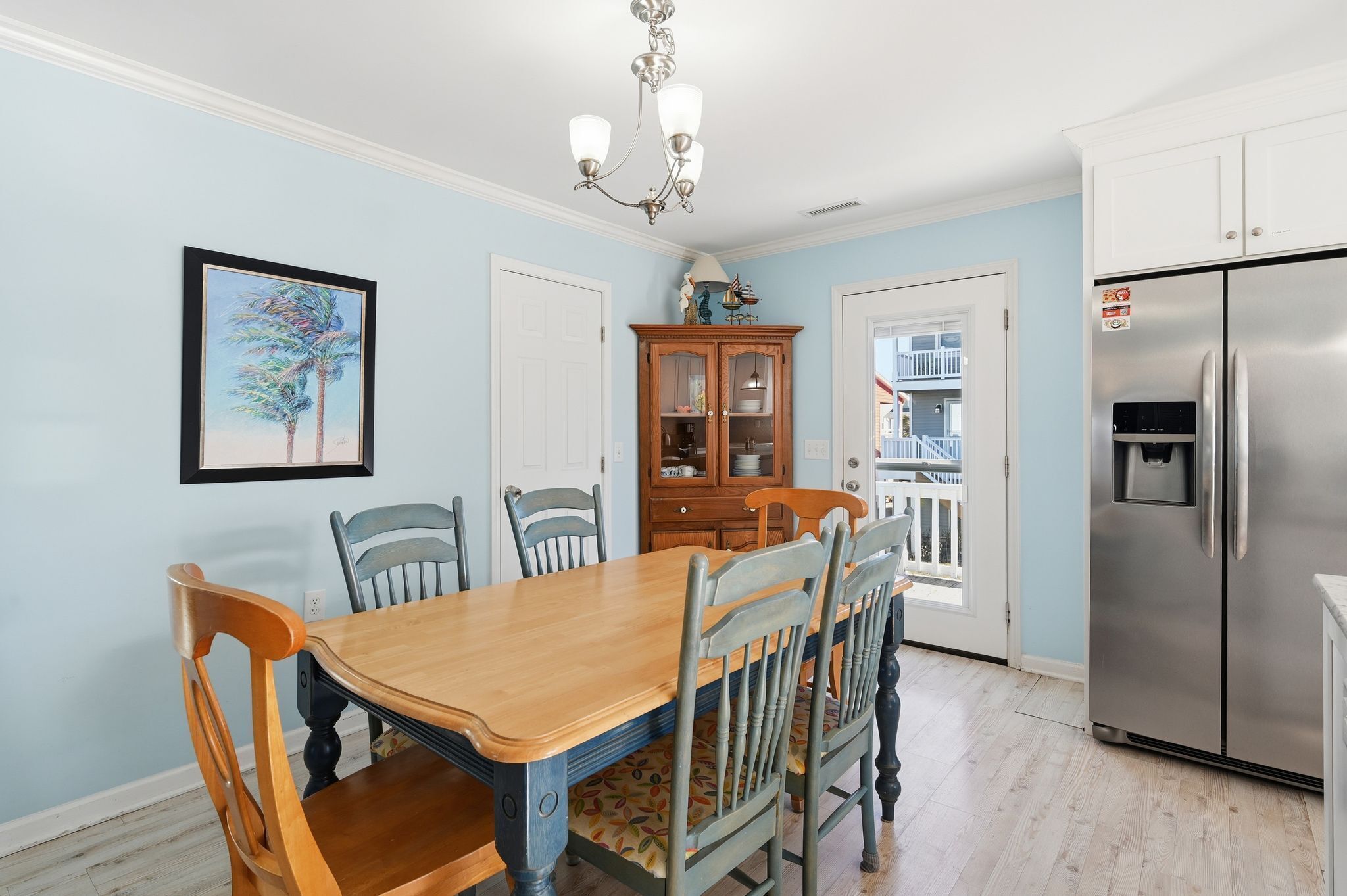 Une salle à manger avec une table et des chaises en bois, un lustre, un réfrigérateur et une porte donnant sur un balcon.