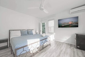 Ein Schlafzimmer mit einem Bett, Deckenventilator, einem am Wand montierten Fernseher und einem Fenster mit Jalousien.