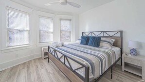 Un dormitorio con una cama, una mesita de noche, un ventilador de techo y ventanas con persianas.
