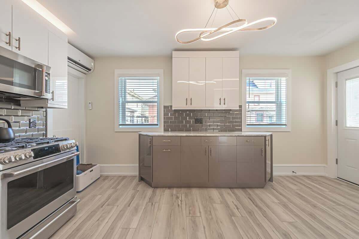 A modern kitchen with a built-in oven, microwave, and gas stove. There are two windows with blinds, and a pendant light hangs above the island.