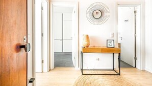A hallway with a wooden door, a round wall decoration, a small wooden table with a vase and picture frame, and a white door with a logo. - Two bedroom apartments close to train station by Lisbon with Sintra (Sintra)