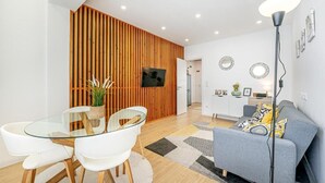A modern living room with a glass dining table, white chairs, a grey sofa, and a wooden accent wall. - Two bedroom apartments close to train station by Lisbon with Sintra (Sintra)