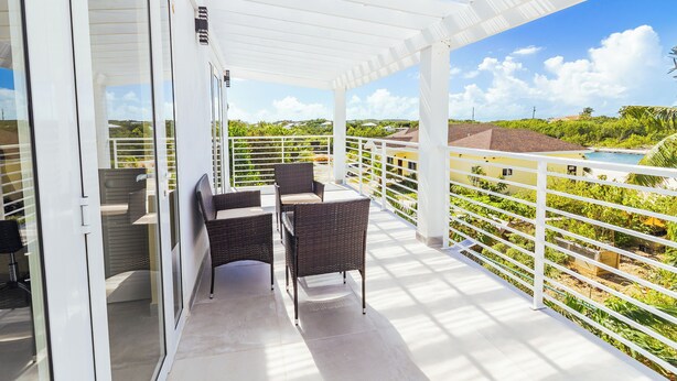 En overdækket balkon med fletmøbler, glasdøre og udsigt til grønt og en swimmingpool.