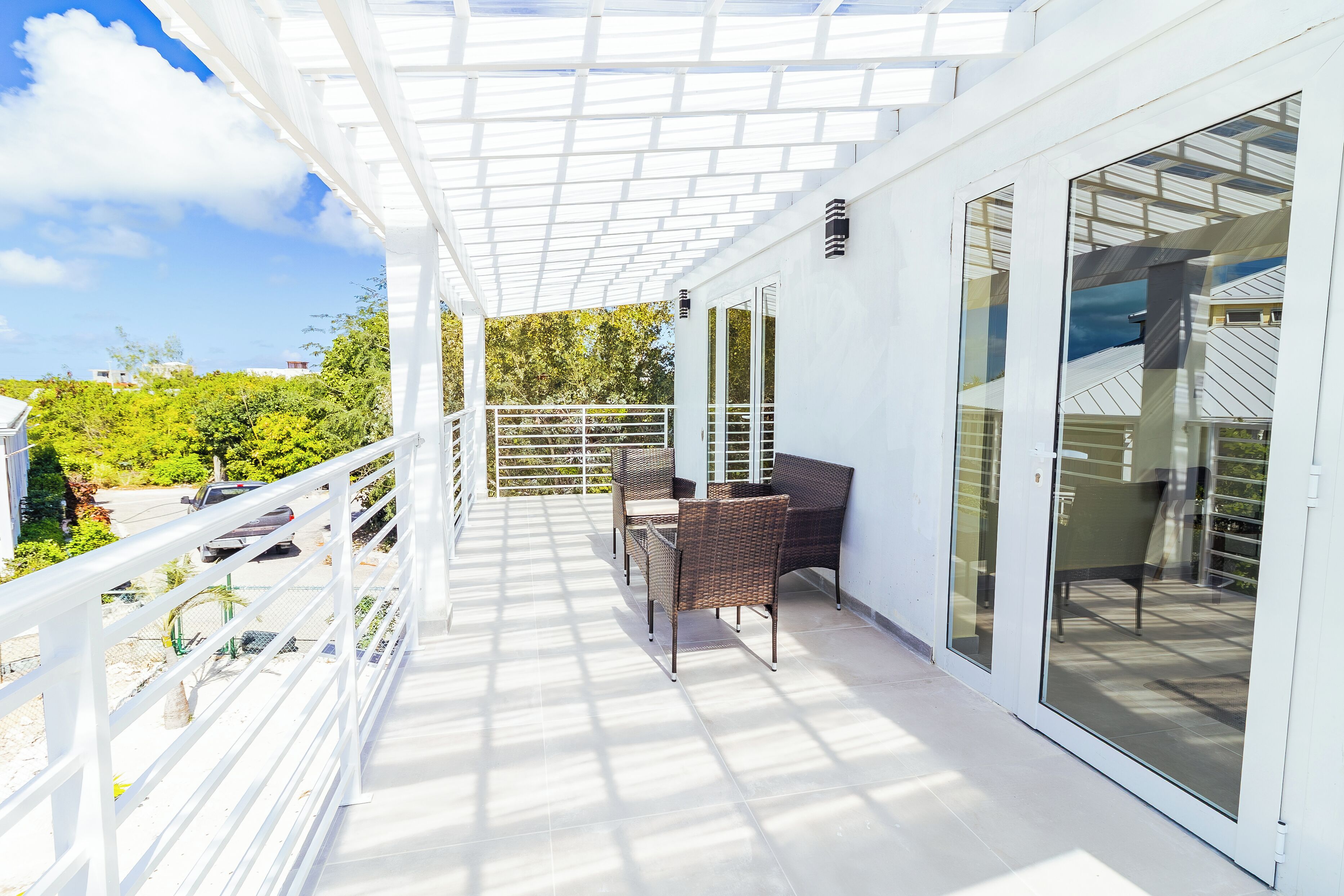A covered balcony with a table and chairs, glass doors, and a view of greenery and buildings.