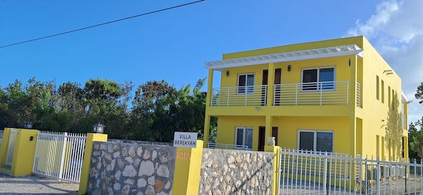 A two-story yellow building with a stone wall entrance, a white fence, and a sign indicating 'VILLA BEREKYAH.'