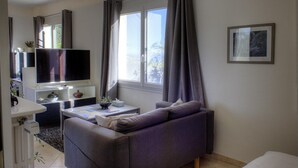 Een moderne woonkamer met een bank, een flatscreen televisie, een klein tafeltje met een plant en een raam met gordijnen.