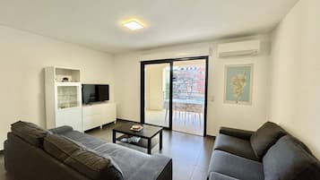Una sala de estar moderna con dos sofás grises, un mueble blanco para el entretenimiento con un televisor, una mesa de centro y un ventanal corredizo que da a un balcón exterior.